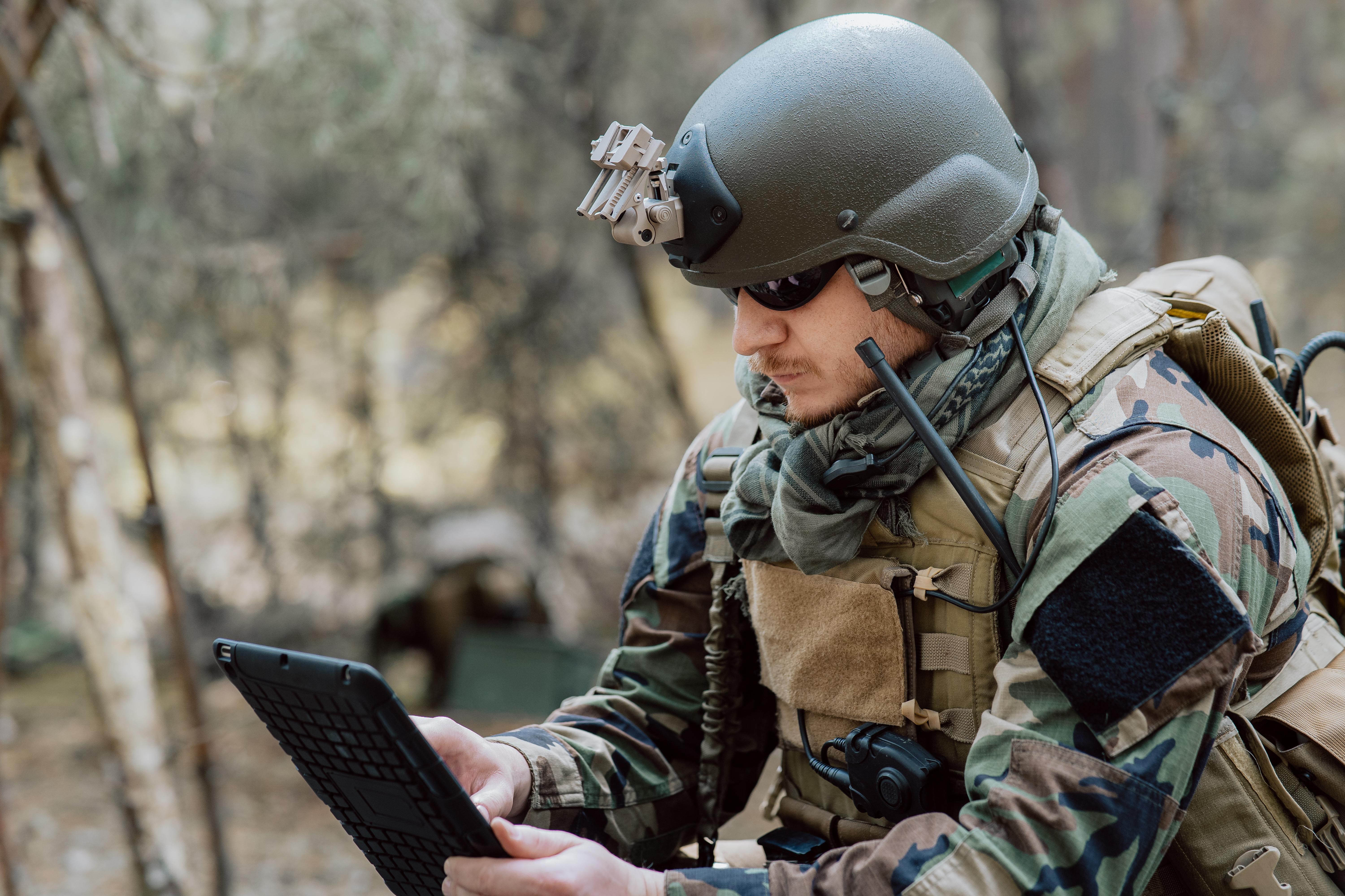 Bearded soldier in uniform sit on military transport crates, analyze data on a tablet and work out tactics at a temporary forest base.